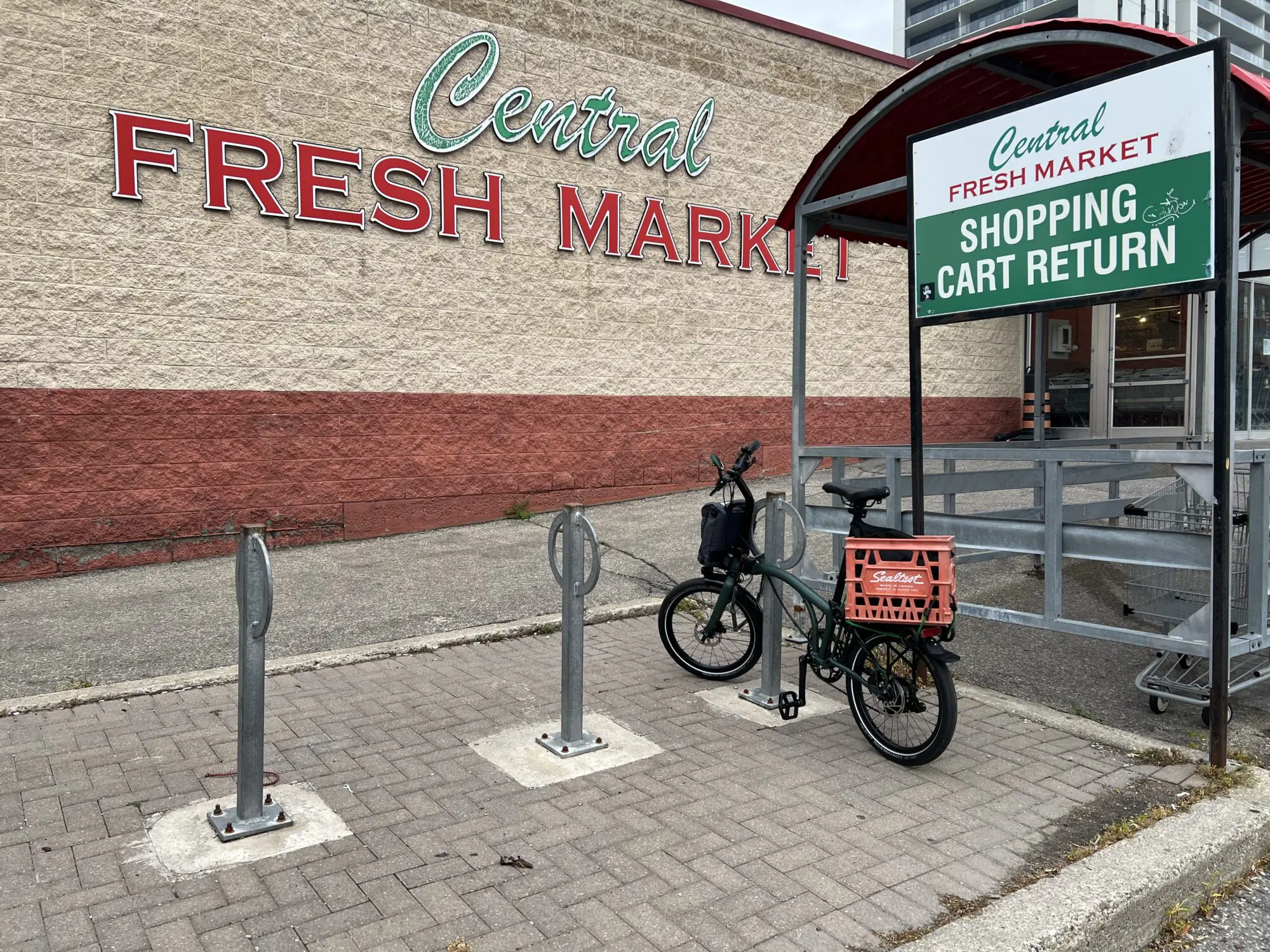A view of a bike parking area, a Brompton locked to a post-and-ring style parking spot. A large crate is mounted on the rerar rack of the bike. In the background thereโs the wall of a building. On the wall the words โCentral Fresh Marketโ are prominent.