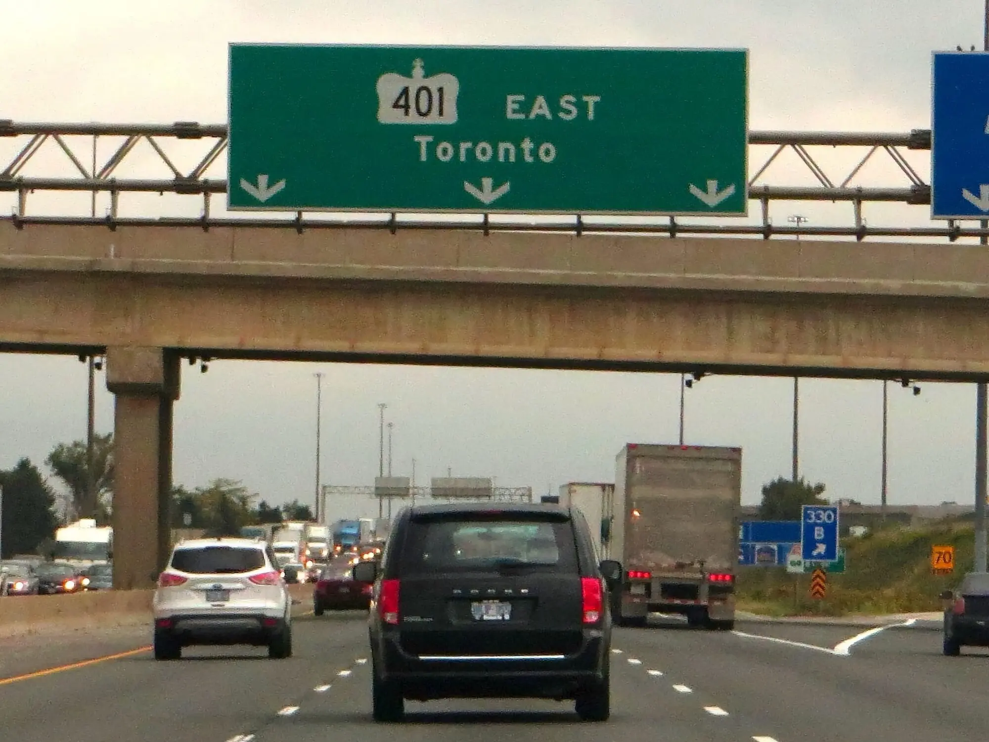 A view of a multilane controlled access highway, looking ahead towards cars in three lanes of traffic. A sign over the lanes reads "401 East. Toronto."