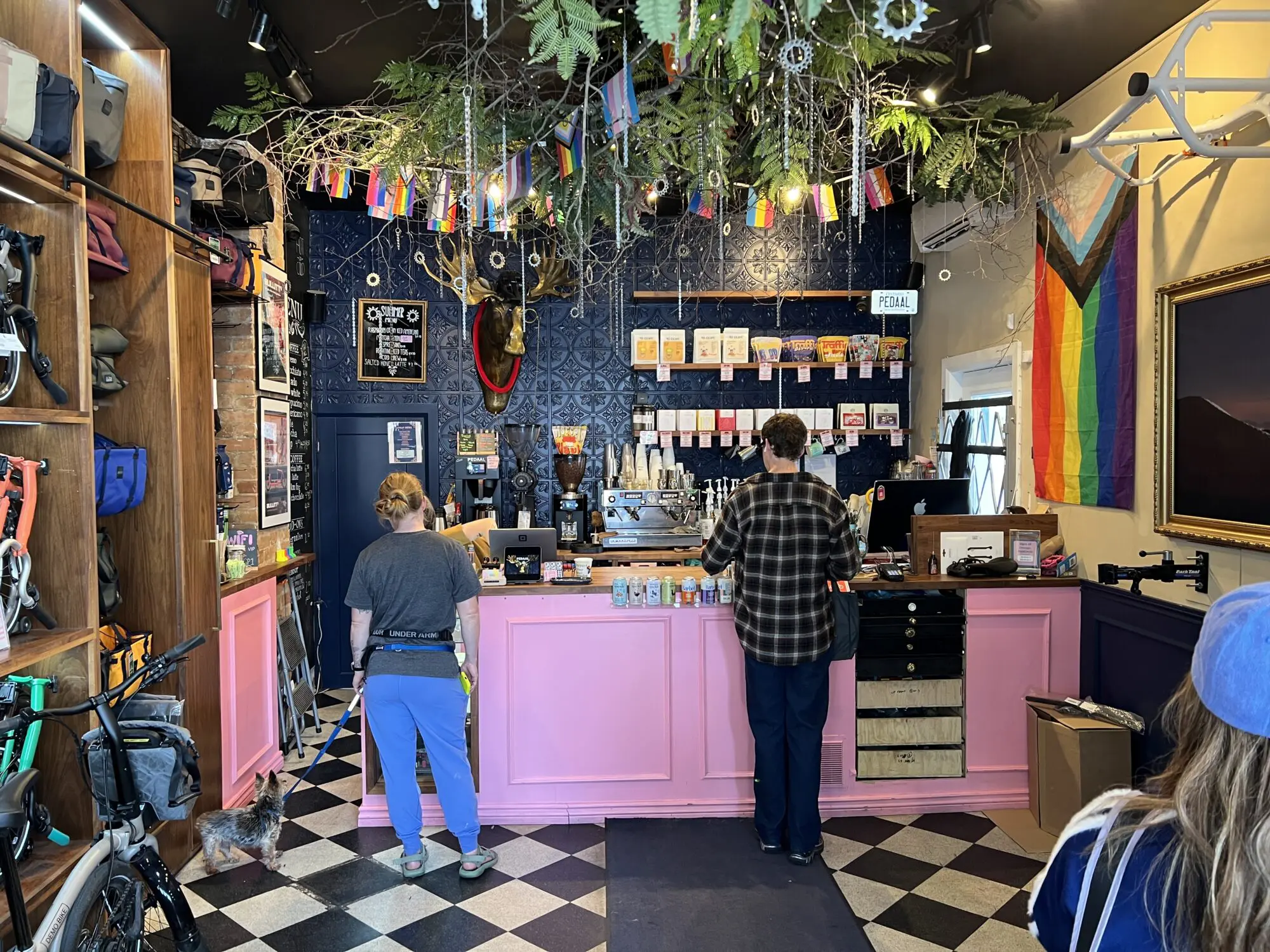 A view into a retail space. At the back there's a counter with various drinks available. There are two people standing at the counter. The ceiling has plants and mini banners hanging down. It's all quite colourful.
