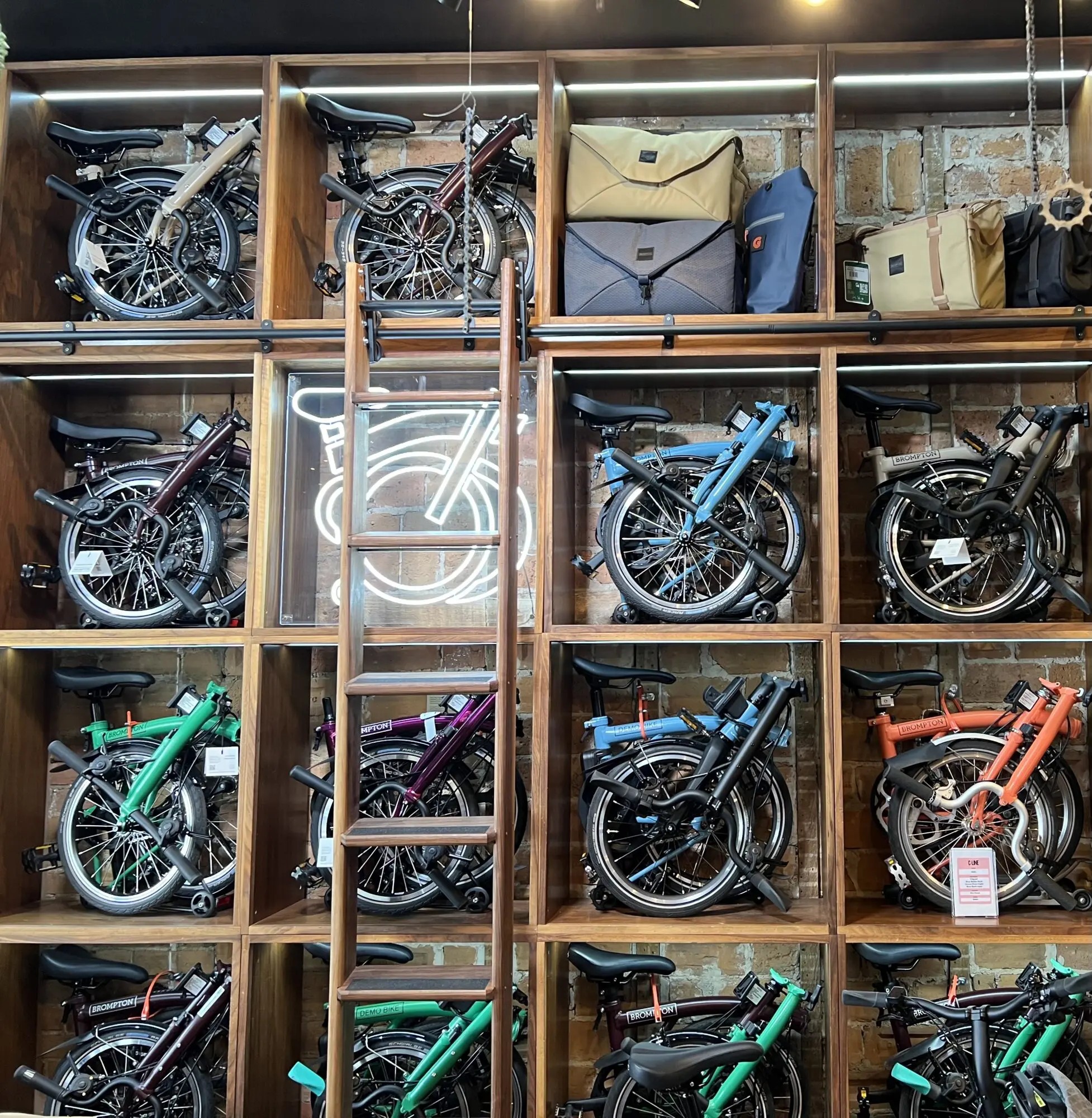 Sixteen cubby hole shelves hold folded Brompton bikes and bags. A ladder rests against the shelves.
