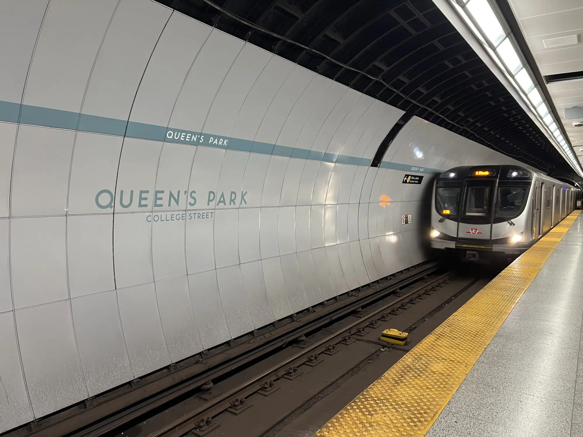 A view of a subway train approaching passenger platform. There's a white tiled wall on the left, with the station name that reads "QUEE'S PARK. COLLEGE STREET."